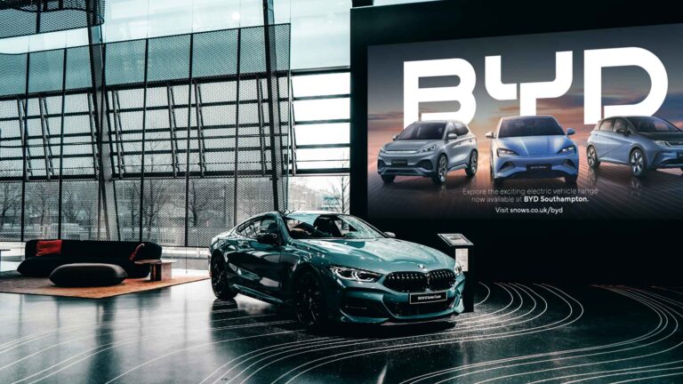 A sleek teal BMW coupe parked in a showroom next to a giant digital advertisement for BYD electric vehicles.